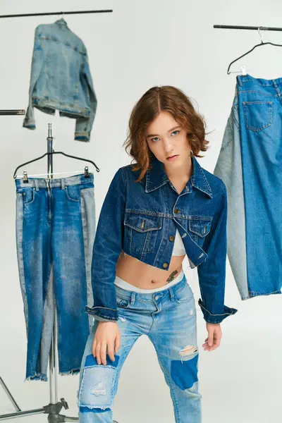 Татуйована молода модель в обрізаному піджаку і джинсах, що стоять серед джинсового одягу на сірому фоні — стокове фото