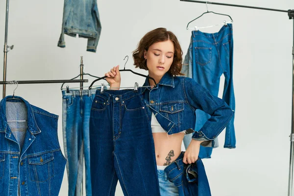 Tattooed young woman in blue cropped jacket holding jeans among denim clothes on grey backdrop — Stock Photo