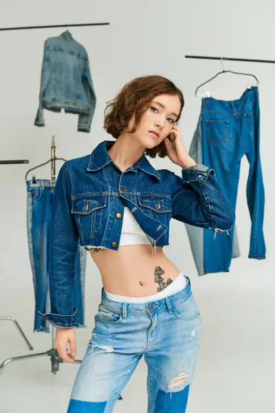 Tattooed young model in cropped jacket and jeans posing among denim clothes on grey backdrop — Stock Photo
