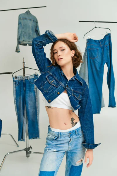 Tätowiertes junges Model in gestutzter Jacke und Jeans posiert mit erhobener Hand zwischen Jeansklamotten auf grau — Stockfoto
