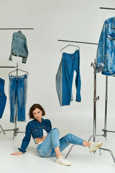 Gen z model in blue cropped jacket and jeans sitting among trendy denim clothes on grey backdrop — Stock Photo