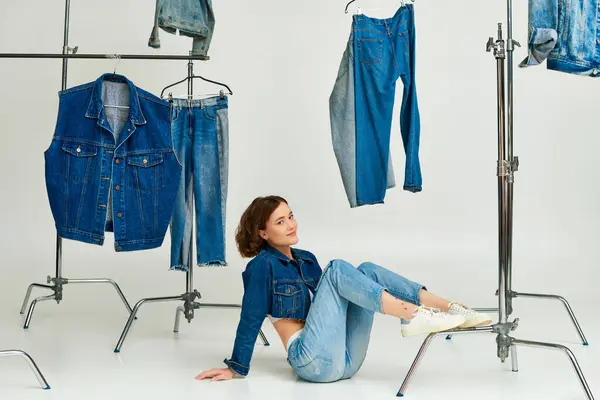 Hübsches junges Model in blauer Kurzjacke und Jeans sitzt inmitten trendiger Jeanskleidung auf grau — Stockfoto