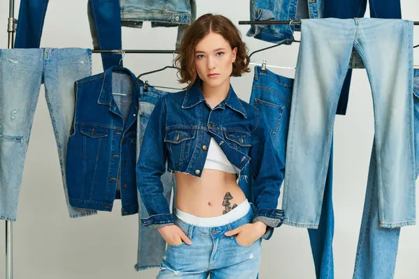 Chic young model in cropped jacket and jeans posing among trendy denim clothes on grey backdrop — Stock Photo