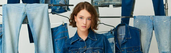 Modelo jovem chique em jaqueta de ganga e jeans posando entre roupas de jeans em pano de fundo cinza, bandeira — Stock Photo