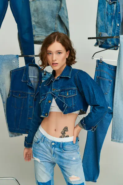 Pretty girl in cropped jacket and jeans posing with hand on hip among trendy denim clothes on grey — Stock Photo