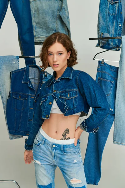 Bella ragazza in giacca ritagliata e jeans in posa con mano sul fianco tra abiti in denim alla moda su grigio — Foto stock