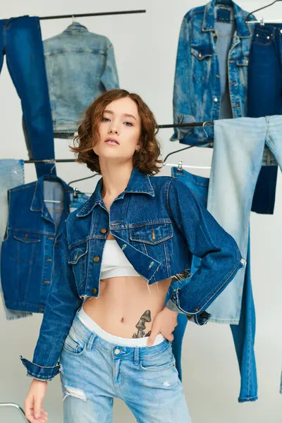 Stylisches Gen-Z-Model in abgeschnittener Jacke und Jeans posiert zwischen Jeanskleidung auf grauem Hintergrund — Stockfoto