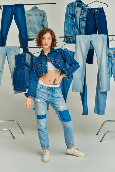 Modèle gen z tendance en veste et jean recadré posant parmi les vêtements en denim sur fond gris — Photo de stock
