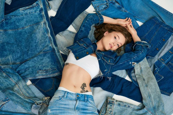 Top view, tattooed young model lying on pile of denim clothes with raised hands, fast fashion — Stock Photo
