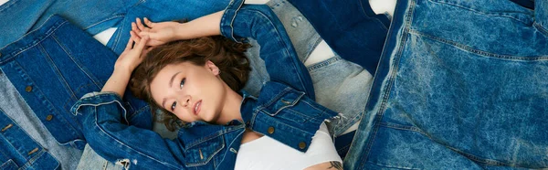 Vista superior, tatuado jovem modelo deitado na pilha de roupas de ganga com as mãos levantadas, bandeira de moda rápida — Fotografia de Stock