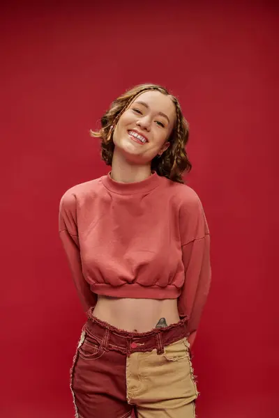 Glückliches Gesicht, tätowiertes Mode-Model posiert in Patchworkhosen und abgeschnittenem Langarm auf rotem Hintergrund — Stockfoto