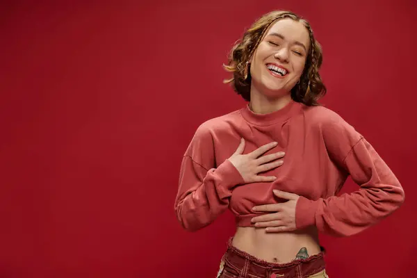 Mujer joven feliz en pantalones patchwork de moda y recortado manga larga riendo sobre fondo rojo - foto de stock