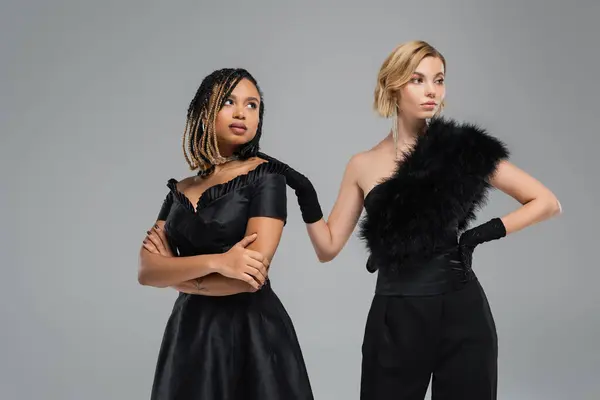 Vielfältige Schönheit, multiethnische Freundinnen im schwarzen eleganten Outfit stehen und wegschauen auf grau — Stockfoto
