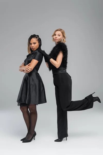 Zwei attraktive multirassische Freundinnen in schwarzer eleganter Kleidung posieren und in die Kamera auf grau schauen — Stockfoto