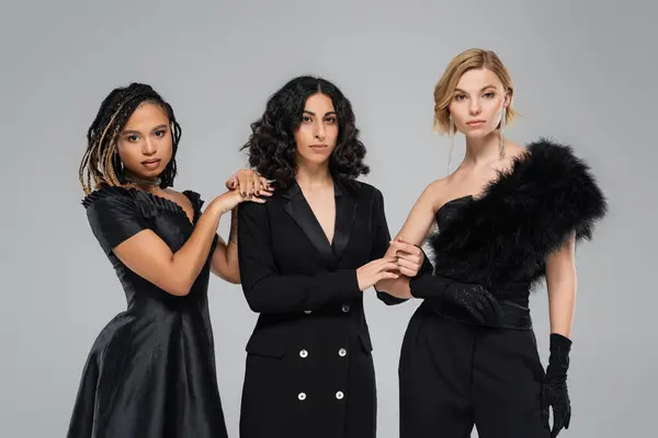 Три стильні жінки в загальному чорному елегантному вбранні на сірому, група сучасних багаторасових подружок — стокове фото