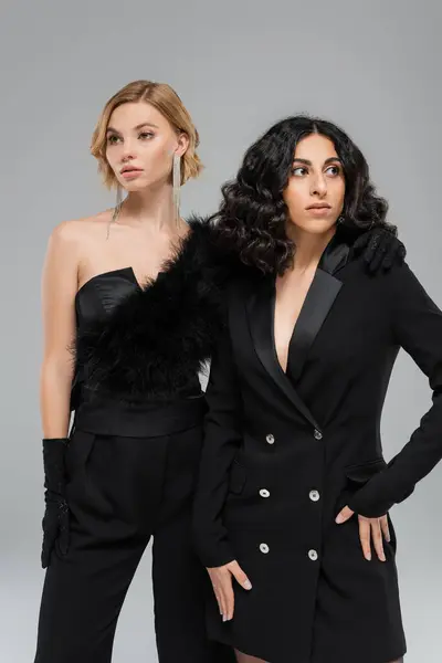 Bruna e bionda fidanzate multirazziali in total black elegante abbigliamento in posa su sfondo grigio — Foto stock