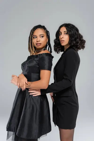 Diversidad y moda, dos amigas multirraciales en ropa elegante negra posando sobre fondo gris - foto de stock