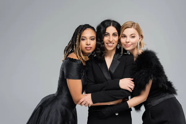 Glückliche multirassische Freundinnen in schwarzen eleganten Outfits, die sich in Grau, Vielfalt und Mode umarmen — Stockfoto