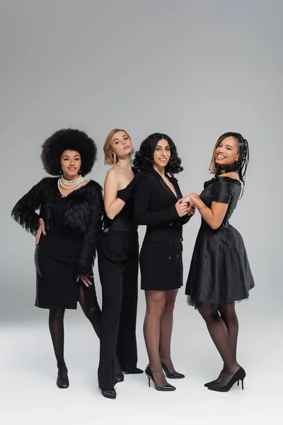 Fröhliche und elegante multirassische Freundinnen in ganz schwarzer Kleidung, die in grau in die Kamera lächeln — Stockfoto