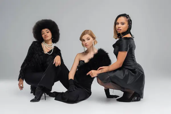 Fashionistas multiétnicos em preto elegante desgaste sentado e olhando para a câmera em cinza, tiro de moda — Fotografia de Stock