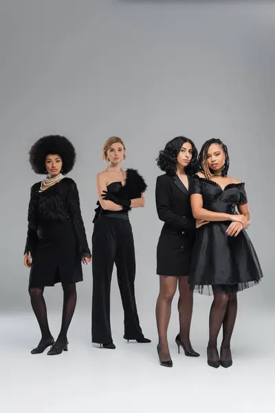 In voller Länge glamouröse modische multirassische Freundinnen in schwarzer Kleidung, die auf grau stehen — Stockfoto