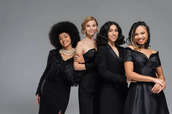 Група радісних багаторасових друзів-жінки в загальному чорному елегантному вбранні, посміхаючись на камеру на сірому — стокове фото