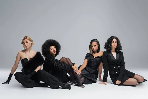 Grupo em elegantes namoradas multirraciais em roupas pretas sentado em cinza, diversidade em alta moda — Fotografia de Stock