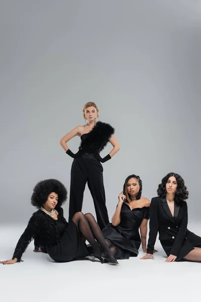 Modelo feminino loira com as mãos nos quadris perto de amigos multiculturais do sexo feminino em preto sentado no cinza — Fotografia de Stock