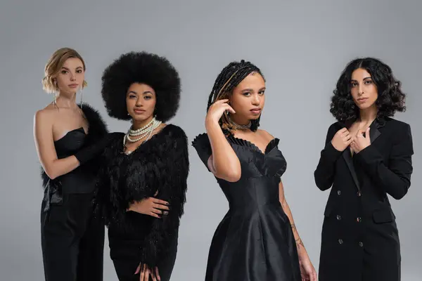 Modernas namoradas multiétnicas em roupas elegantes pretas olhando para a câmera em cinza, beleza diversificada — Fotografia de Stock