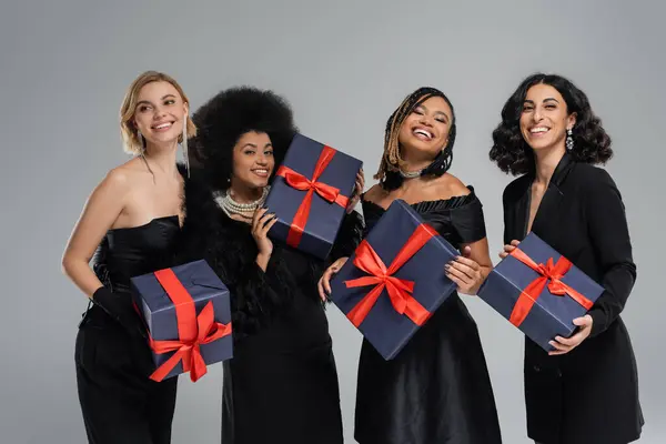Grupo de mujeres multiétnicas alegres en traje elegante negro celebración de regalos de vacaciones en gris - foto de stock