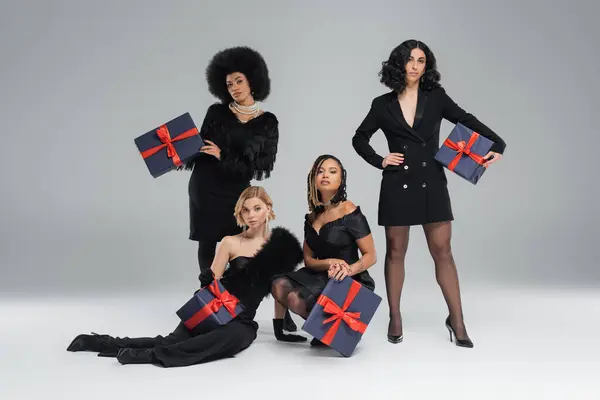 Modelos de moda multiculturales posando en ropa elegante negro con cajas de regalo festivas en gris - foto de stock