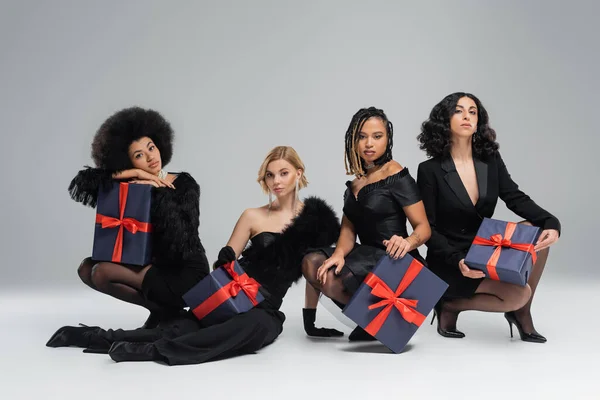 Gruppe von verschiedenen multirassischen Freundinnen in insgesamt schwarzer stylischer Kleidung sitzt mit Geschenkboxen auf grau — Stockfoto