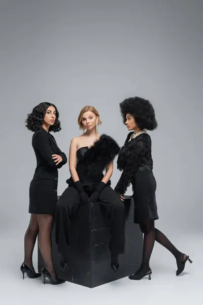 Elegante blonde Frau sitzt auf schwarzem Würfel neben multiethnischen Freundinnen in dunkler Kleidung auf grau — Stockfoto