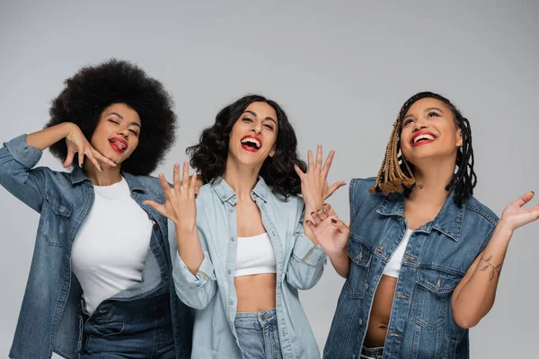 Overjoyed multiracial girlfriends in blue denim clothes gesturing on grey, friendship and happiness — Stock Photo