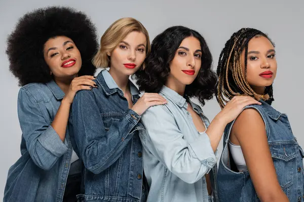 Multiracial girlfriends in jeans wear holding hands on shoulders of each other and smiling at camera — Stock Photo