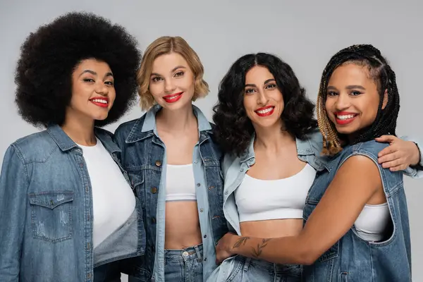 Group of smiley multicultural girlfriends in denim clothes looking at camera on grey, diversity — Stock Photo