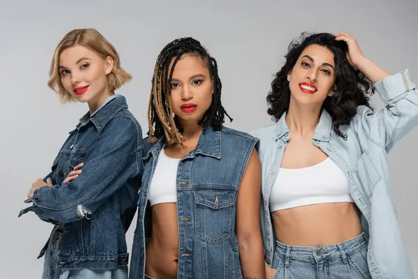 Fashionable multiracial girlfriends in blue denim clothes looking at camera on grey, diverse beauty — Stock Photo