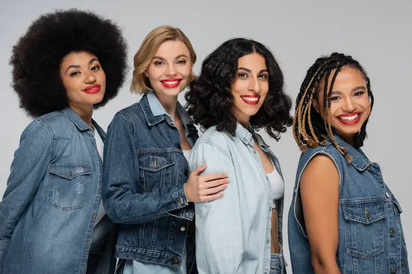Joyful multicultural female models in casual denim clothes smiling at camera on grey, diverse beauty — Stock Photo