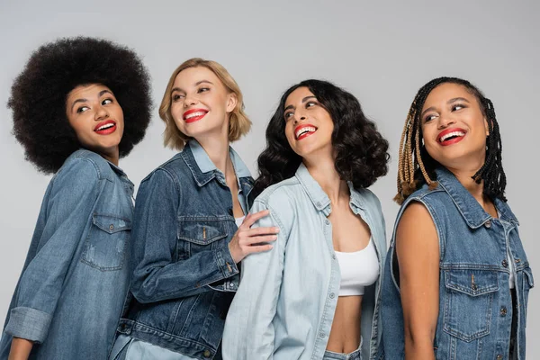 Cheerful multiethnic girlfriends in casual denim clothes looking away on grey, diversity and fashion — Stock Photo