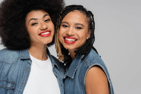 Jeunes et heureuses copines afro-américaines en denim bleu regardant la caméra sur gris — Photo de stock