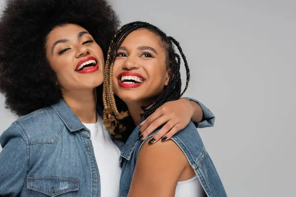 Excited and stylish african american woman embracing girlfriend on grey backdrop, denim style — Stock Photo