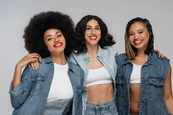 Young and fashionable multiracial female friends in blue denim outfit smiling at camera on grey — Stock Photo