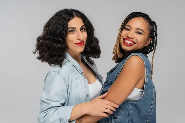 Two carefree multiracial girlfriends in stylish denim attire smiling at camera on grey, togetherness — Stock Photo