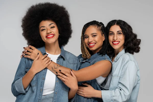 Stylish and smiley multiethnic girlfriends in blue jeans wear looking at camera on grey, diversity — Stock Photo