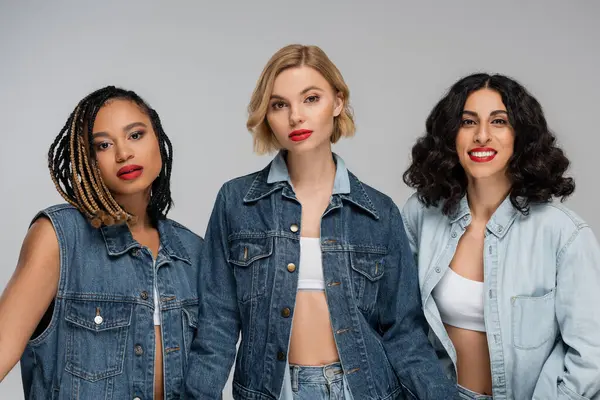 Three attractive multiracial women in denim clothes looking at camera on grey, group portrait — Stock Photo