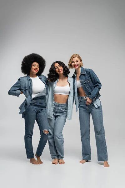 Full length of barefoot multiracial girlfriends in blue denim clothes posing on grey backdrop — Stock Photo