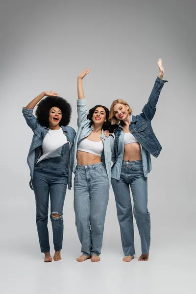 Full length of joyful barefoot multiethnic girlfriends in blue denim wear waving hands on grey — Stock Photo