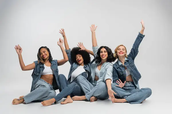 Excited and barefoot multiracial girlfriends in denim clothes sitting and waving hands grey — Stock Photo