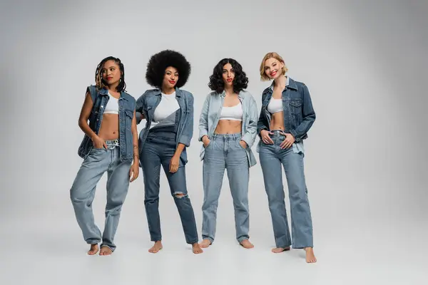 Босоніж багатонаціональні подружки в стильному джинсовому одязі, що стоїть босоніж на сірому, повнометражному — стокове фото