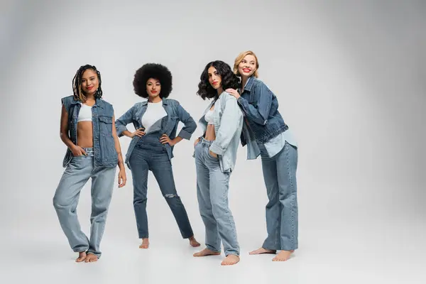 Confident multiracial girlfriends in trendy denim street wear standing barefoot on grey, full length — Stock Photo